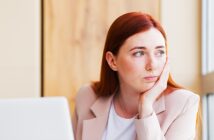 A woman with long red hair and a light pink blazer is sitting at a desk, resting her chin in her hand. She is looking thoughtfully out the window next to her, with an open laptop in front of her.