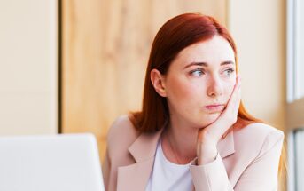 A woman with long red hair and a light pink blazer is sitting at a desk, resting her chin in her hand. She is looking thoughtfully out the window next to her, with an open laptop in front of her.