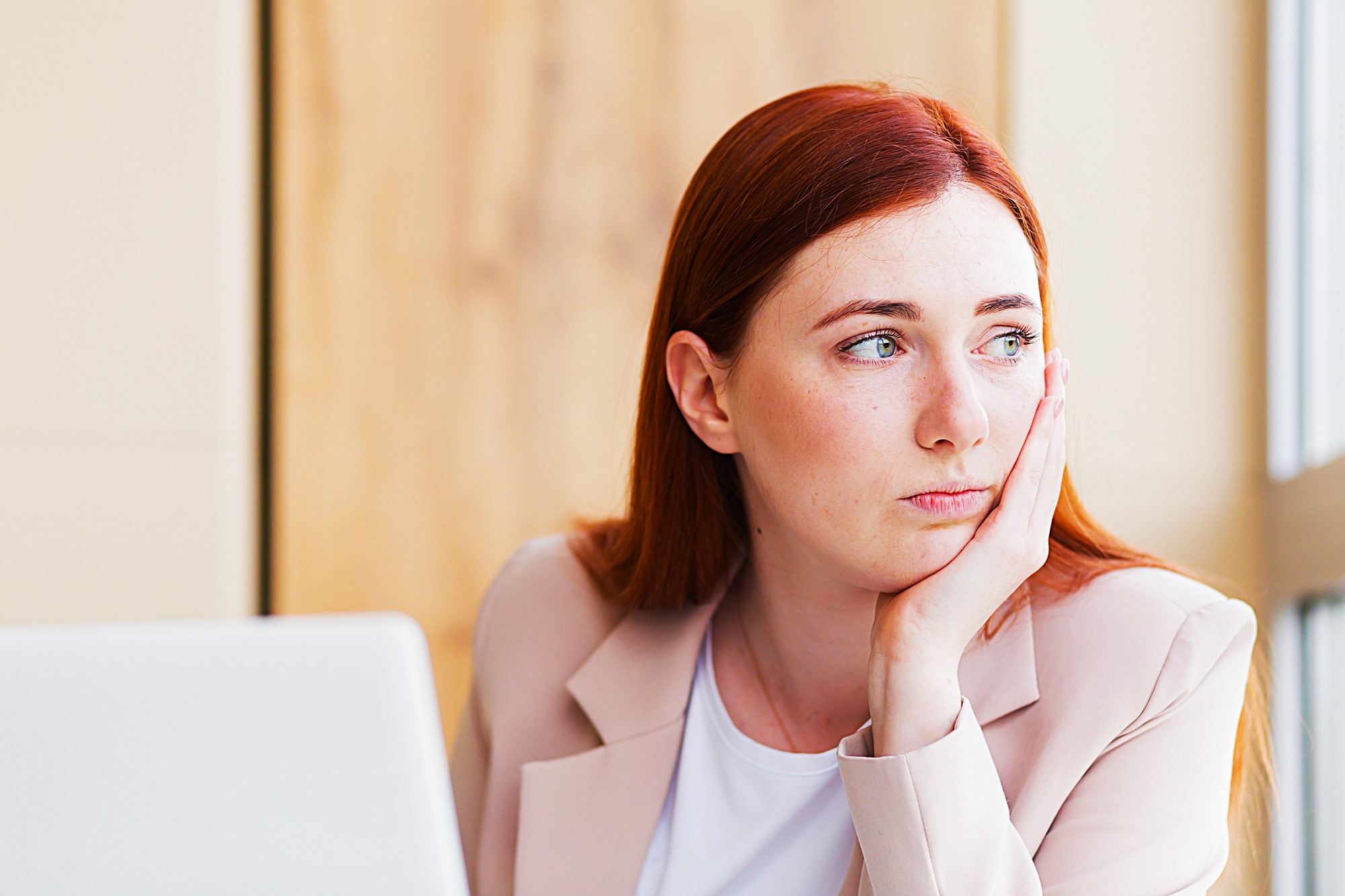 A woman with long red hair and a light pink blazer is sitting at a desk, resting her chin in her hand. She is looking thoughtfully out the window next to her, with an open laptop in front of her.