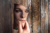 A person holds a broken mirror reflecting their face, appearing through a hole in a weathered wooden surface. The image conveys a sense of introspection or mystery.