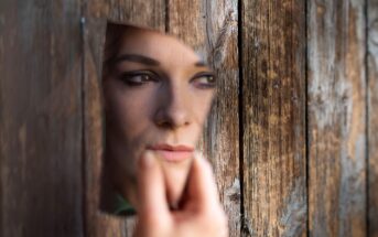 A person holds a broken mirror reflecting their face, appearing through a hole in a weathered wooden surface. The image conveys a sense of introspection or mystery.