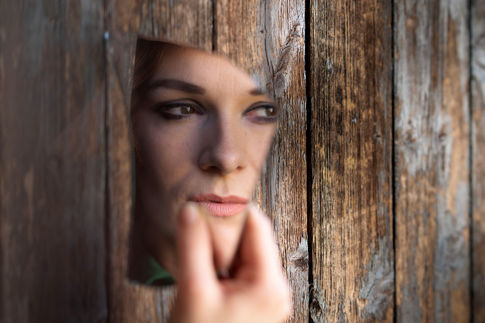 A person holds a broken mirror reflecting their face, appearing through a hole in a weathered wooden surface. The image conveys a sense of introspection or mystery.