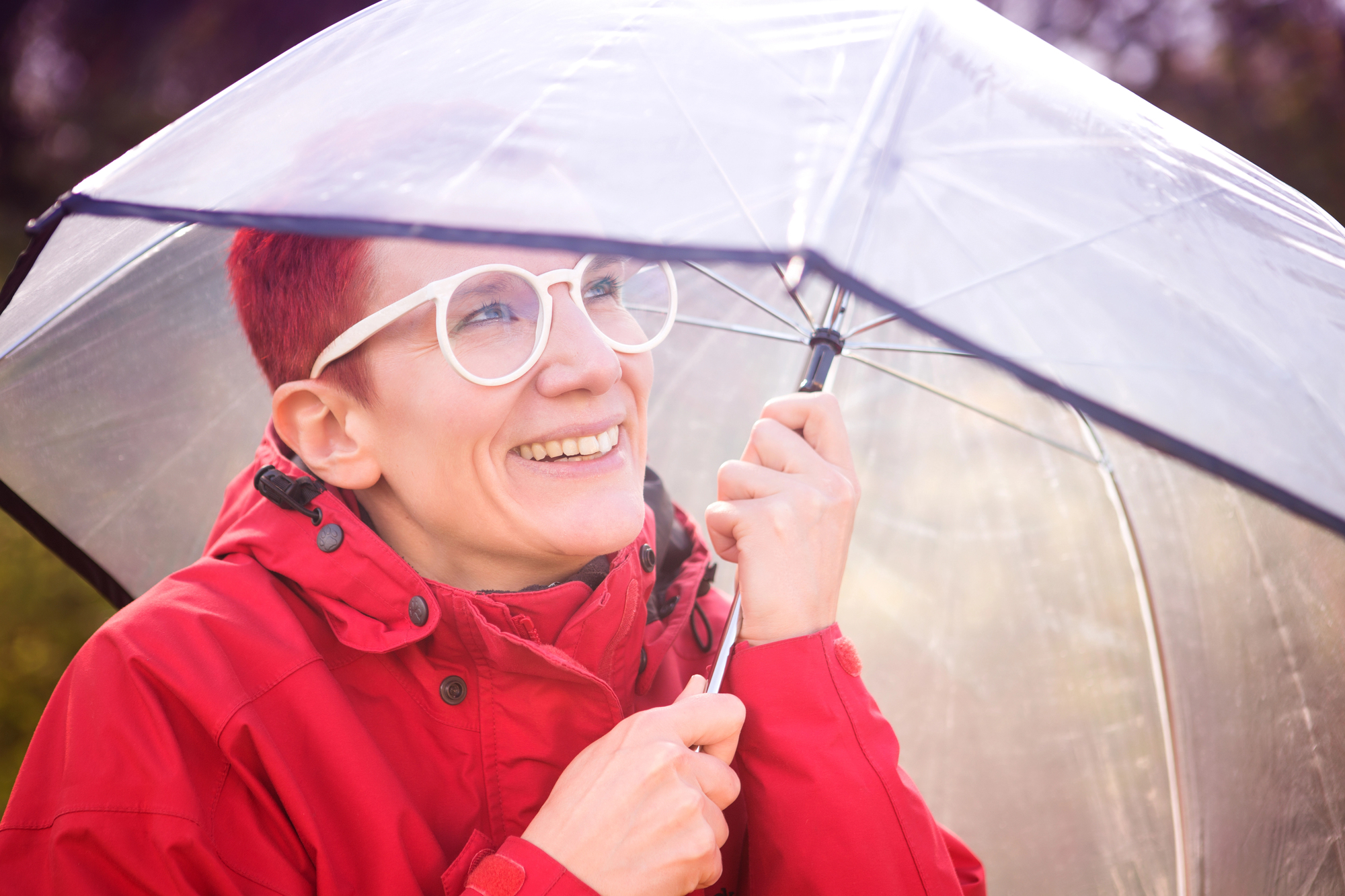 A person with short red hair and glasses smiles under a clear umbrella, wearing a bright red raincoat. The background is softly blurred, suggesting a rainy day in a natural setting.