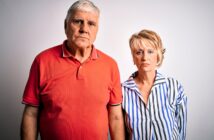 An older couple stands against a plain white background. The man is wearing a red polo shirt and the woman is in a striped blouse. Both have a serious expression.