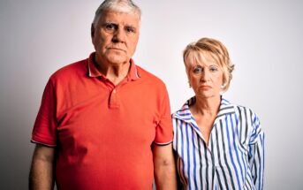 An older couple stands against a plain white background. The man is wearing a red polo shirt and the woman is in a striped blouse. Both have a serious expression.