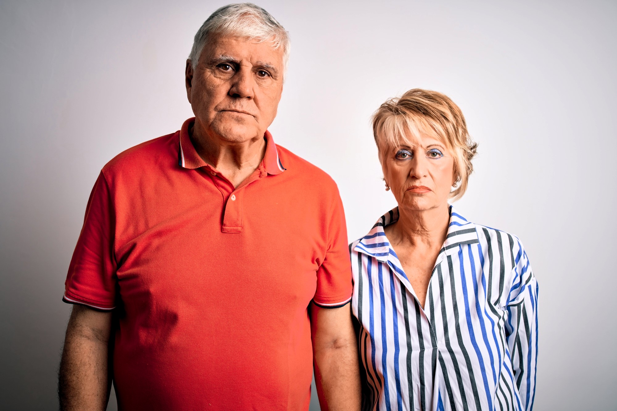 An older couple stands against a plain white background. The man is wearing a red polo shirt and the woman is in a striped blouse. Both have a serious expression.