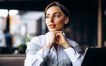 A woman in a white shirt sits at a table in a cafe with her chin resting on her hands. She looks thoughtfully to the side, with a laptop and a cup on the table in front of her. The background is softly blurred.