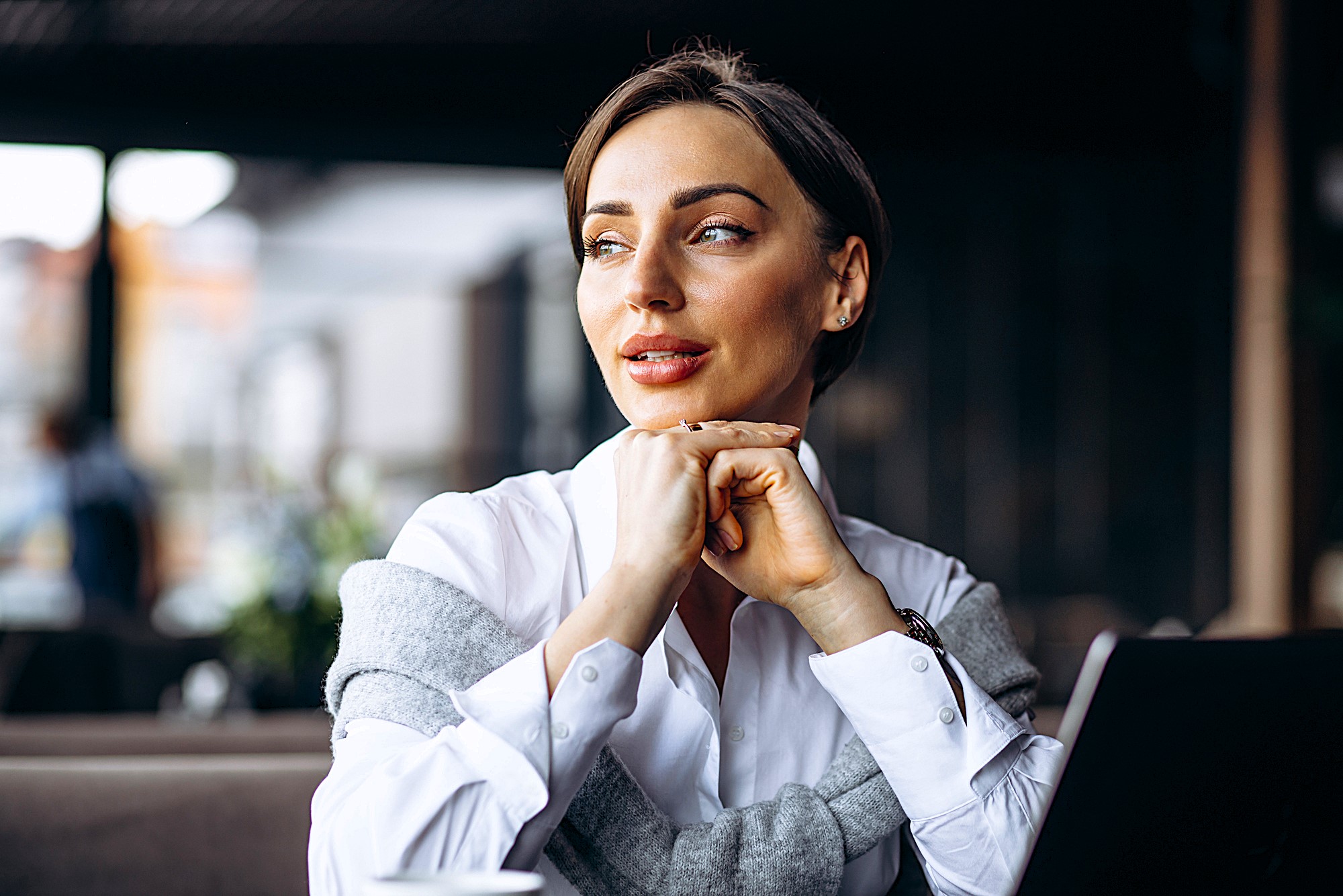 A woman in a white shirt sits at a table in a cafe with her chin resting on her hands. She looks thoughtfully to the side, with a laptop and a cup on the table in front of her. The background is softly blurred.