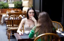 Two women are sitting at an outdoor café. One woman, facing the camera, has long brown hair, looks surprised, and has her hand over her mouth. The other woman, with her back to the camera, is wearing a green top. Both have drinks in front of them.