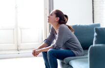 A woman with a ponytail sits on a teal sofa, gazing thoughtfully upwards. Her elbows rest on her knees, and she wears a gray long-sleeve shirt and jeans. The room is softly lit, with a large window in the background.