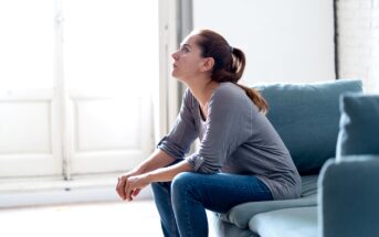 A woman with a ponytail sits on a teal sofa, gazing thoughtfully upwards. Her elbows rest on her knees, and she wears a gray long-sleeve shirt and jeans. The room is softly lit, with a large window in the background.