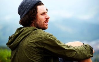 A man wearing a gray beanie and green jacket sits outdoors, looking contemplatively into the distance. His curly hair is visible, and a light drizzle has left water droplets on his jacket. The background is a hazy landscape.