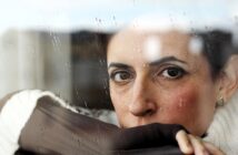 A woman with short hair looks through a rain-speckled window, resting her chin on her hand wrapped in a brown fabric. She wears a white sweater and has a thoughtful expression.