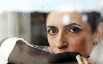 A woman with short hair looks through a rain-speckled window, resting her chin on her hand wrapped in a brown fabric. She wears a white sweater and has a thoughtful expression.