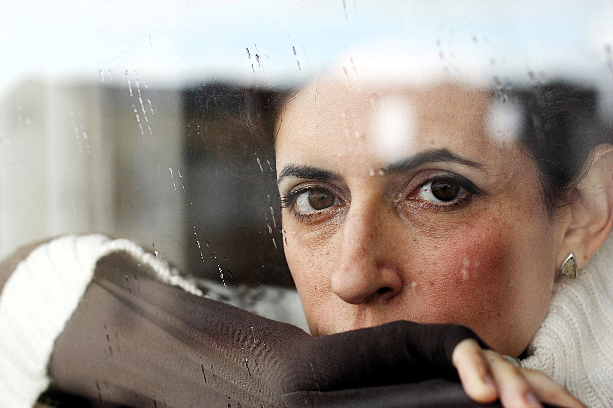 A woman with short hair looks through a rain-speckled window, resting her chin on her hand wrapped in a brown fabric. She wears a white sweater and has a thoughtful expression.