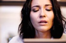 A person with shoulder-length dark hair looks downward with a thoughtful expression. The background is softly blurred, suggesting an indoor setting.