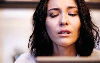 A person with shoulder-length dark hair looks downward with a thoughtful expression. The background is softly blurred, suggesting an indoor setting.