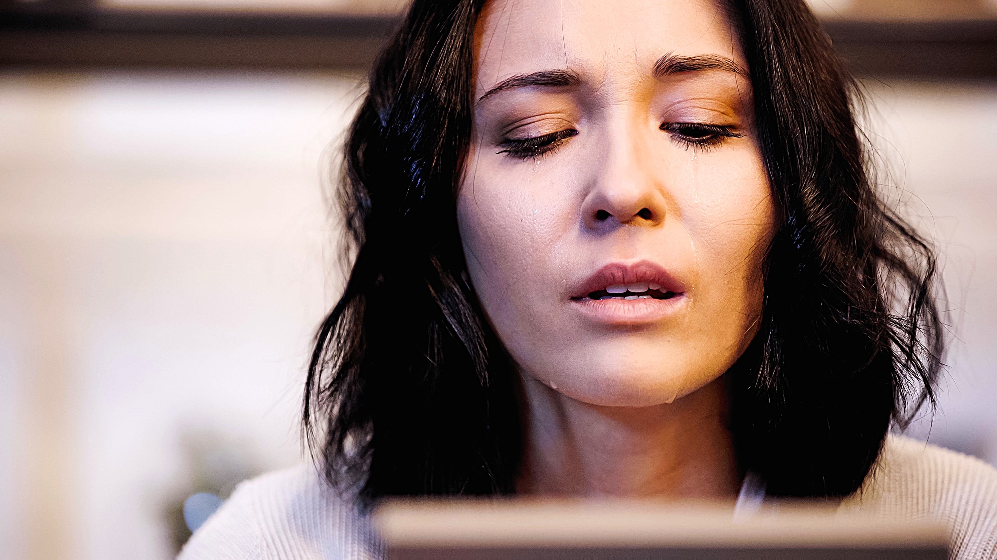 A person with shoulder-length dark hair looks downward with a thoughtful expression. The background is softly blurred, suggesting an indoor setting.