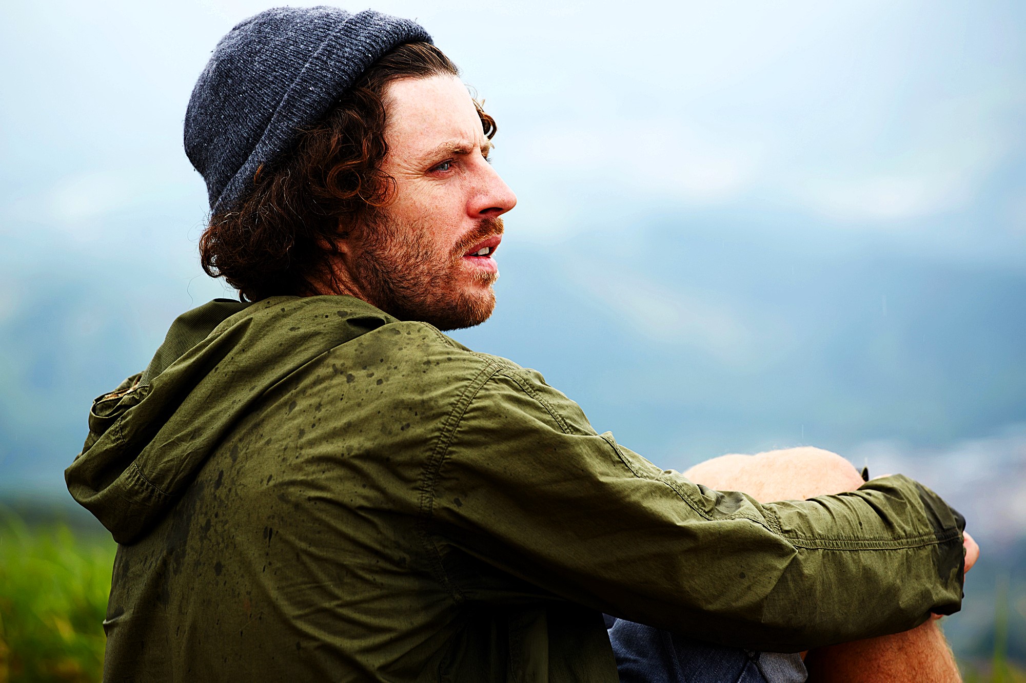 A man wearing a gray beanie and green jacket sits outdoors, looking contemplatively into the distance. His curly hair is visible, and a light drizzle has left water droplets on his jacket. The background is a hazy landscape.