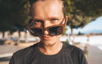 A person with light brown hair is wearing sunglasses, looking at the camera with a slight smile. They are outdoors on a sunny day, and there are trees and a walkway in the blurred background.