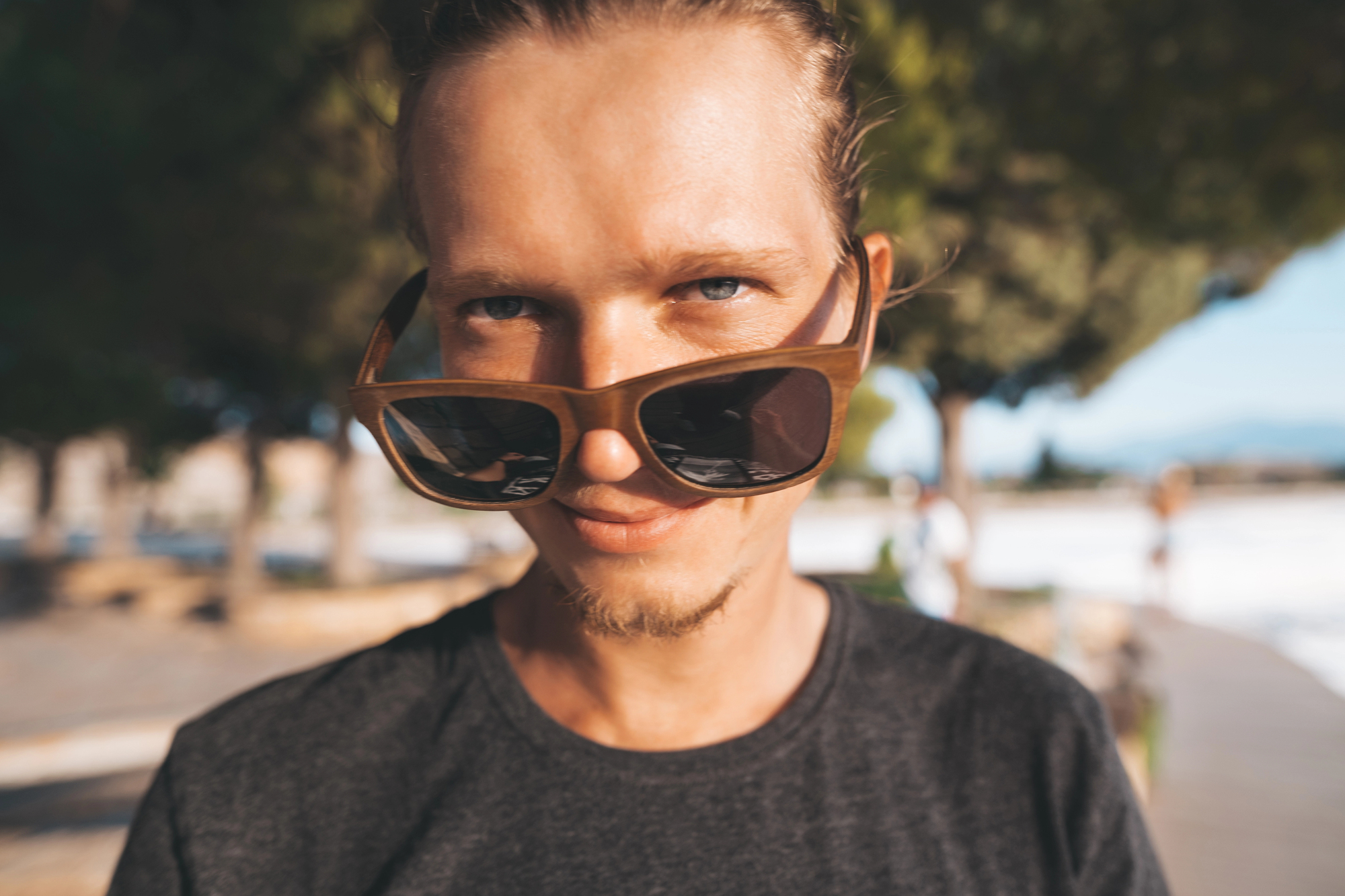 A person with light brown hair is wearing sunglasses, looking at the camera with a slight smile. They are outdoors on a sunny day, and there are trees and a walkway in the blurred background.