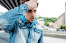 A man in a denim jacket and blue hoodie stands outdoors, looking concerned or thoughtful. He runs a hand through his hair, with buildings and trees blurred in the background, suggesting an urban setting.