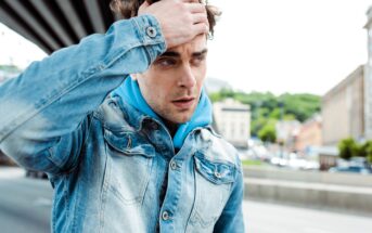 A man in a denim jacket and blue hoodie stands outdoors, looking concerned or thoughtful. He runs a hand through his hair, with buildings and trees blurred in the background, suggesting an urban setting.