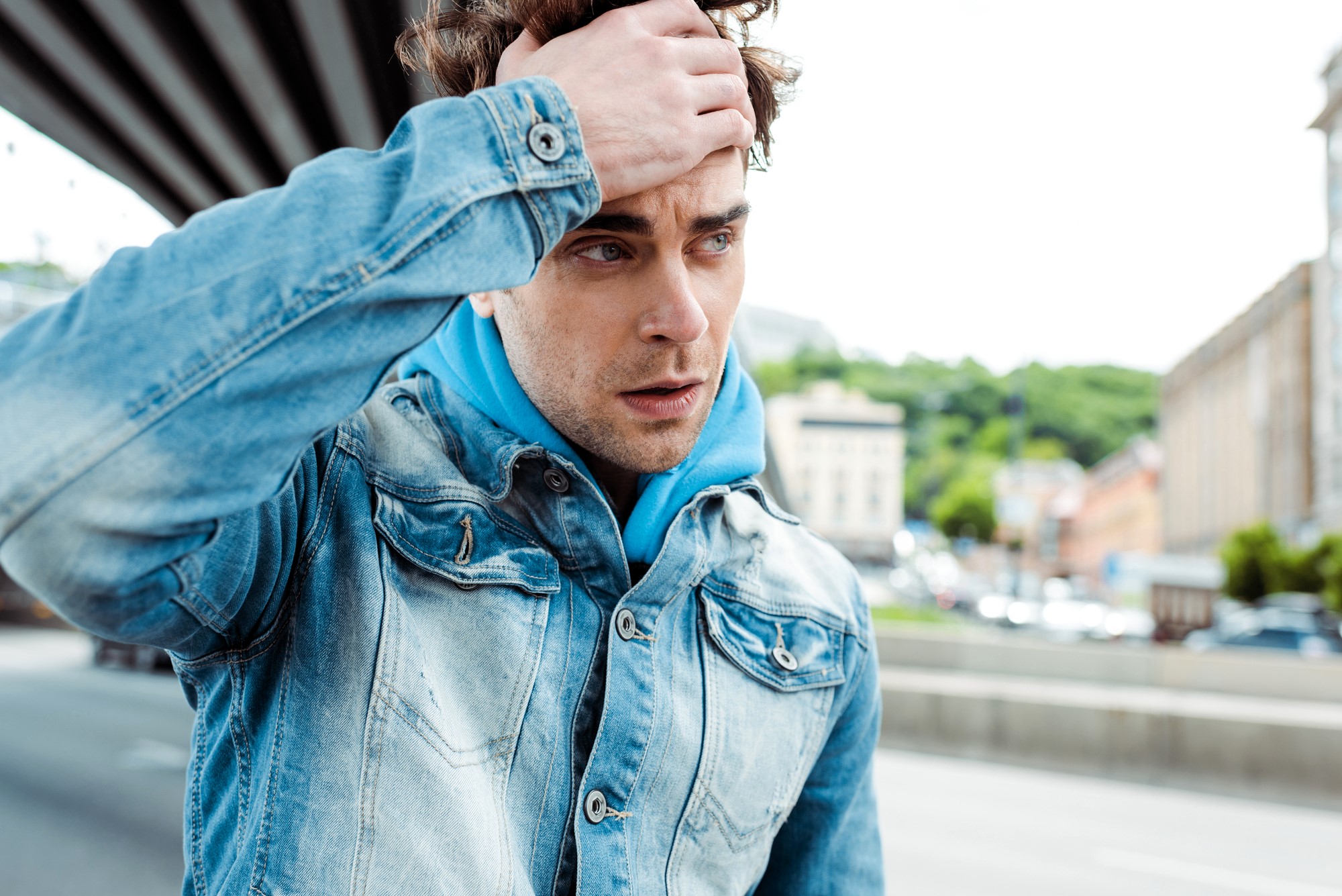 A man in a denim jacket and blue hoodie stands outdoors, looking concerned or thoughtful. He runs a hand through his hair, with buildings and trees blurred in the background, suggesting an urban setting.