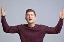 A person in a dark sweater stands with eyes looking upwards and arms raised, appearing puzzled or questioning, against a plain gray background.