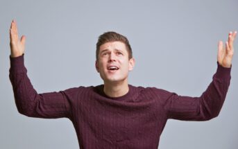 A person in a dark sweater stands with eyes looking upwards and arms raised, appearing puzzled or questioning, against a plain gray background.