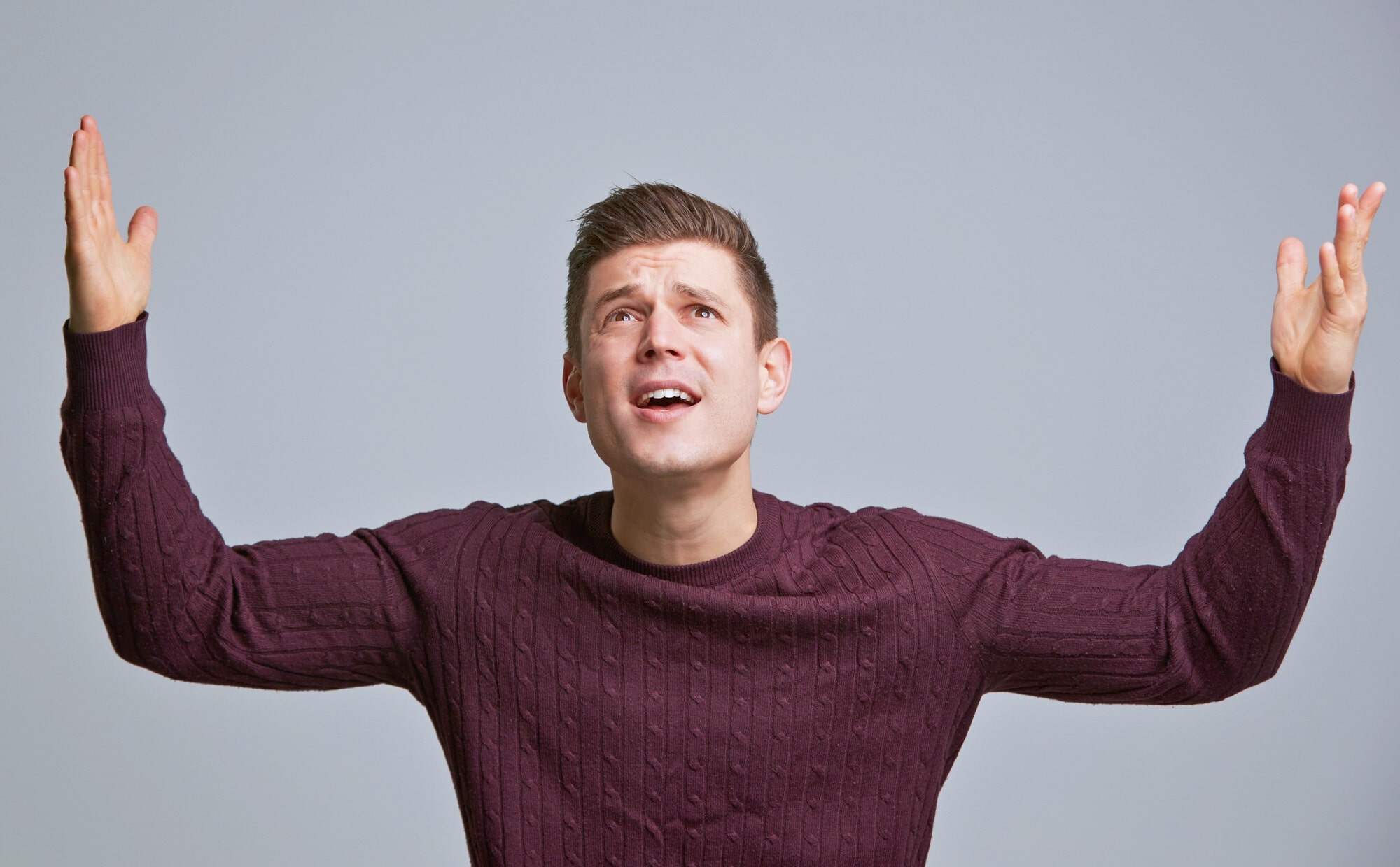 A person in a dark sweater stands with eyes looking upwards and arms raised, appearing puzzled or questioning, against a plain gray background.