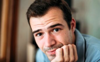 A person with short dark hair rests their chin on their hand, looking intently at the camera. They are wearing a light blue shirt. The background is softly blurred.