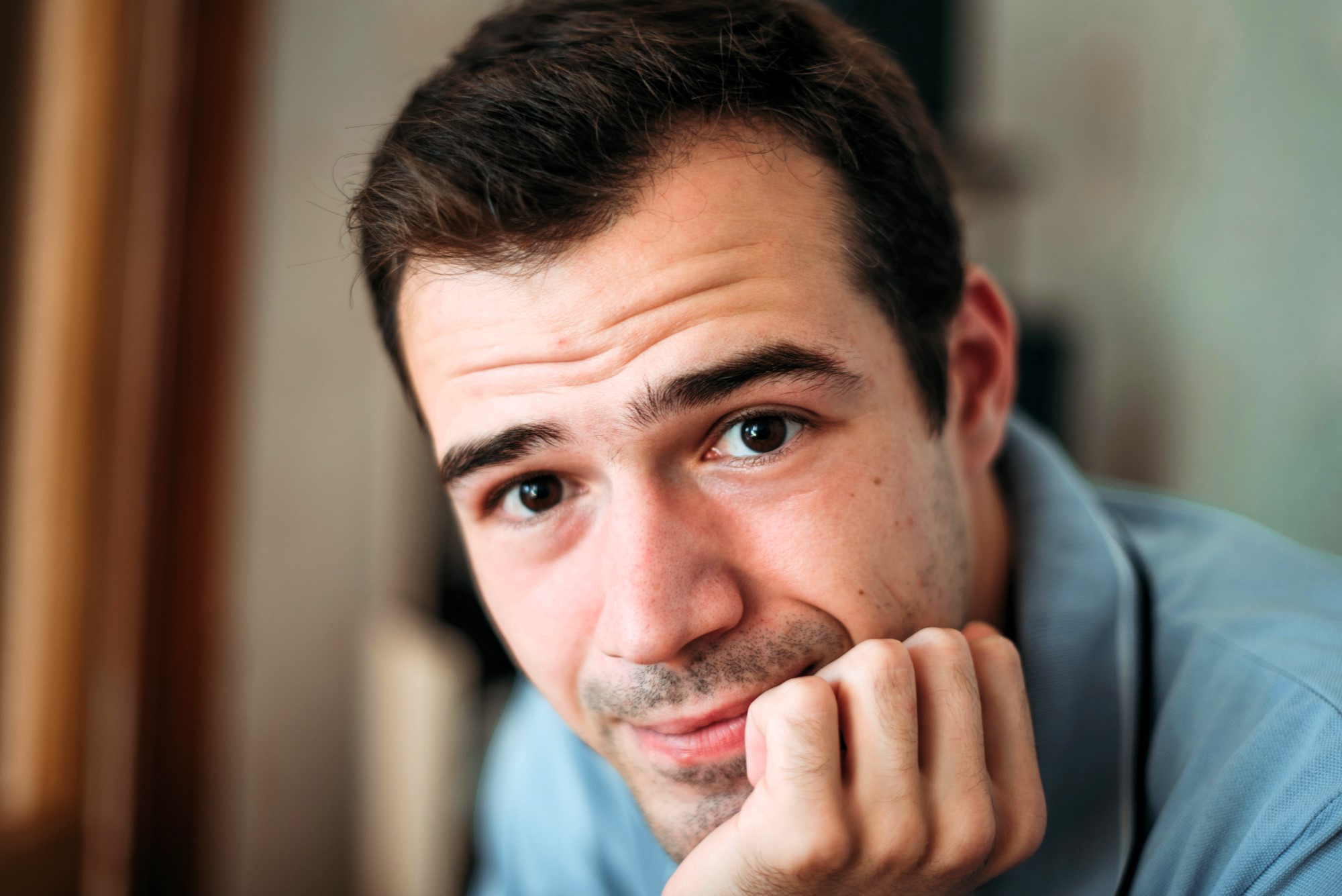 A person with short dark hair rests their chin on their hand, looking intently at the camera. They are wearing a light blue shirt. The background is softly blurred.