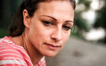 Woman with light brown hair and a contemplative expression, wearing a pink and white striped shirt, sits outdoors. The background is blurred with greenery, suggesting a natural setting.