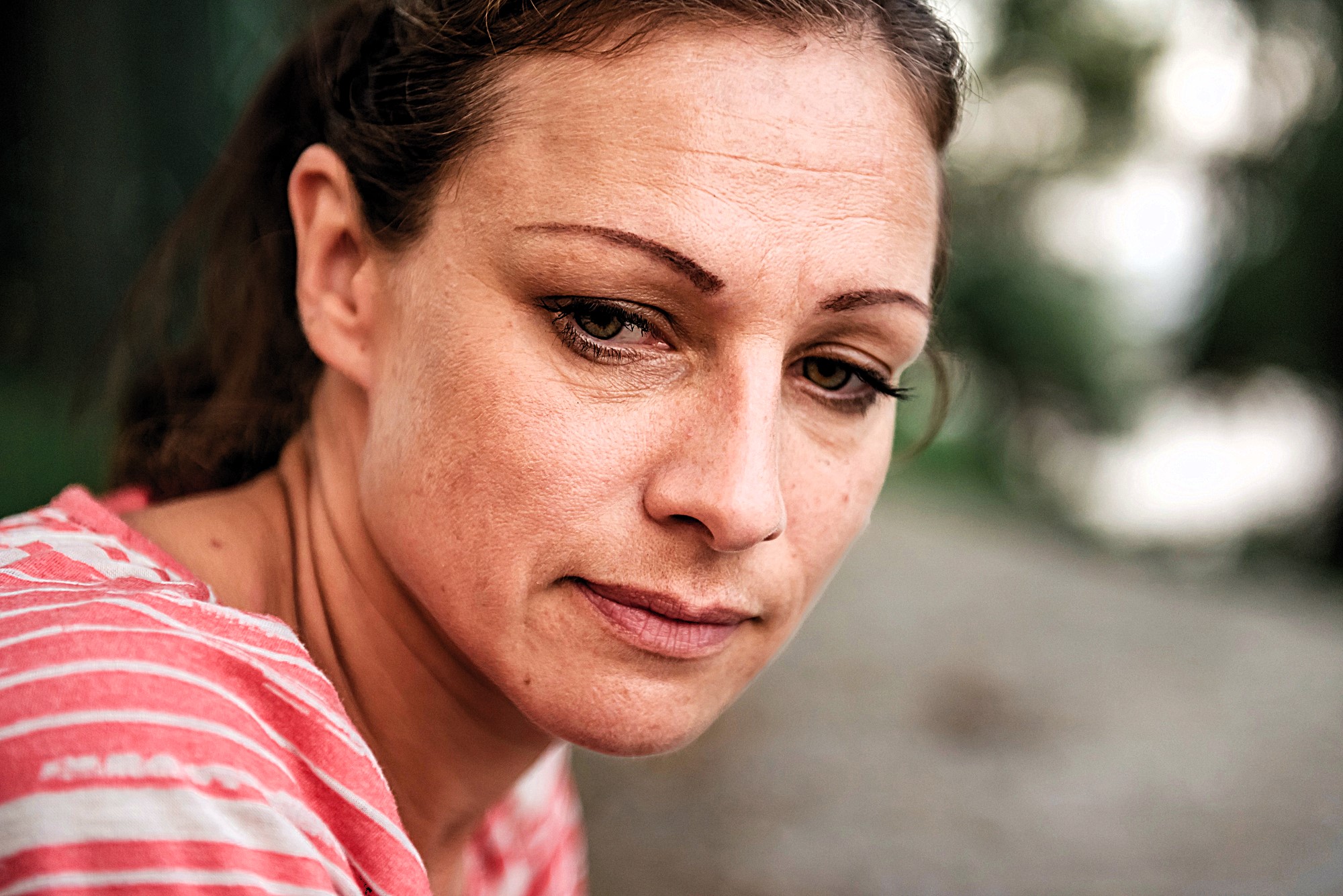 Woman with light brown hair and a contemplative expression, wearing a pink and white striped shirt, sits outdoors. The background is blurred with greenery, suggesting a natural setting.