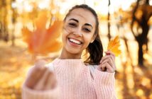 A smiling woman in a pink sweater holds a yellow autumn leaf towards the camera. The background shows a sunlit park with trees and fallen leaves, capturing a warm, autumn day.