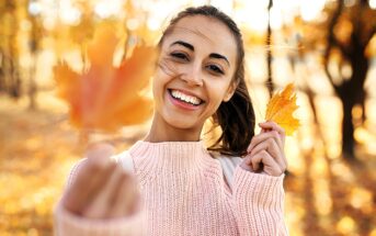 A smiling woman in a pink sweater holds a yellow autumn leaf towards the camera. The background shows a sunlit park with trees and fallen leaves, capturing a warm, autumn day.