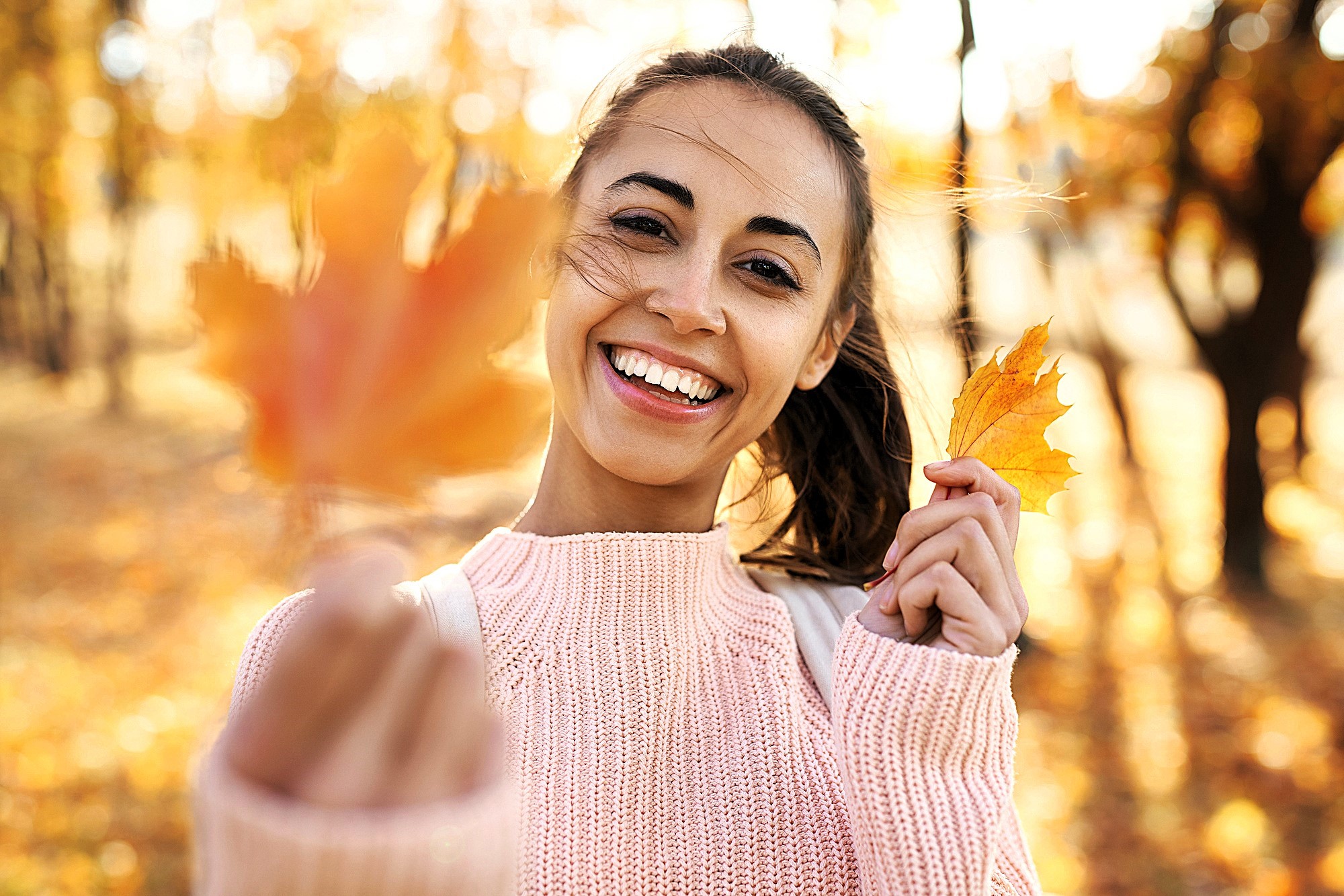 A smiling woman in a pink sweater holds a yellow autumn leaf towards the camera. The background shows a sunlit park with trees and fallen leaves, capturing a warm, autumn day.