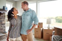 A couple stands in a living room filled with moving boxes, looking at each other and smiling. The room has large windows letting in natural light and a fireplace on one side. They appear to be in the process of moving into a new home.