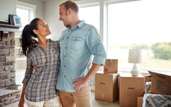 A couple stands in a living room filled with moving boxes, looking at each other and smiling. The room has large windows letting in natural light and a fireplace on one side. They appear to be in the process of moving into a new home.