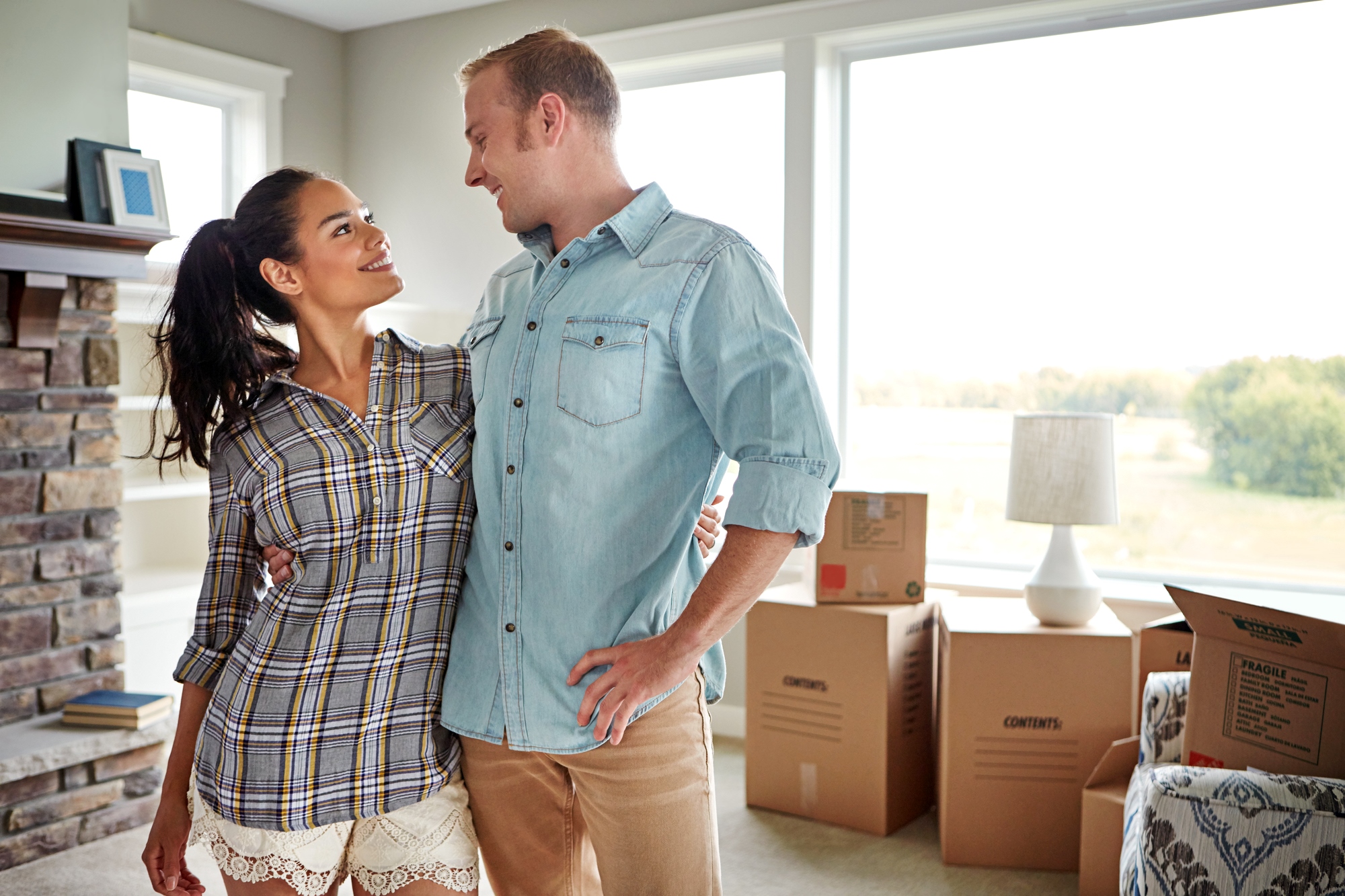 A couple stands in a living room filled with moving boxes, looking at each other and smiling. The room has large windows letting in natural light and a fireplace on one side. They appear to be in the process of moving into a new home.