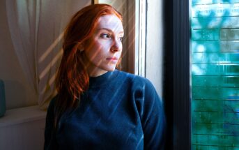 A person with long red hair wearing a dark sweater looks pensively out of a window. Sunlight filters through blinds, casting striped shadows on their face. The window reflects a mix of green foliage and sky.