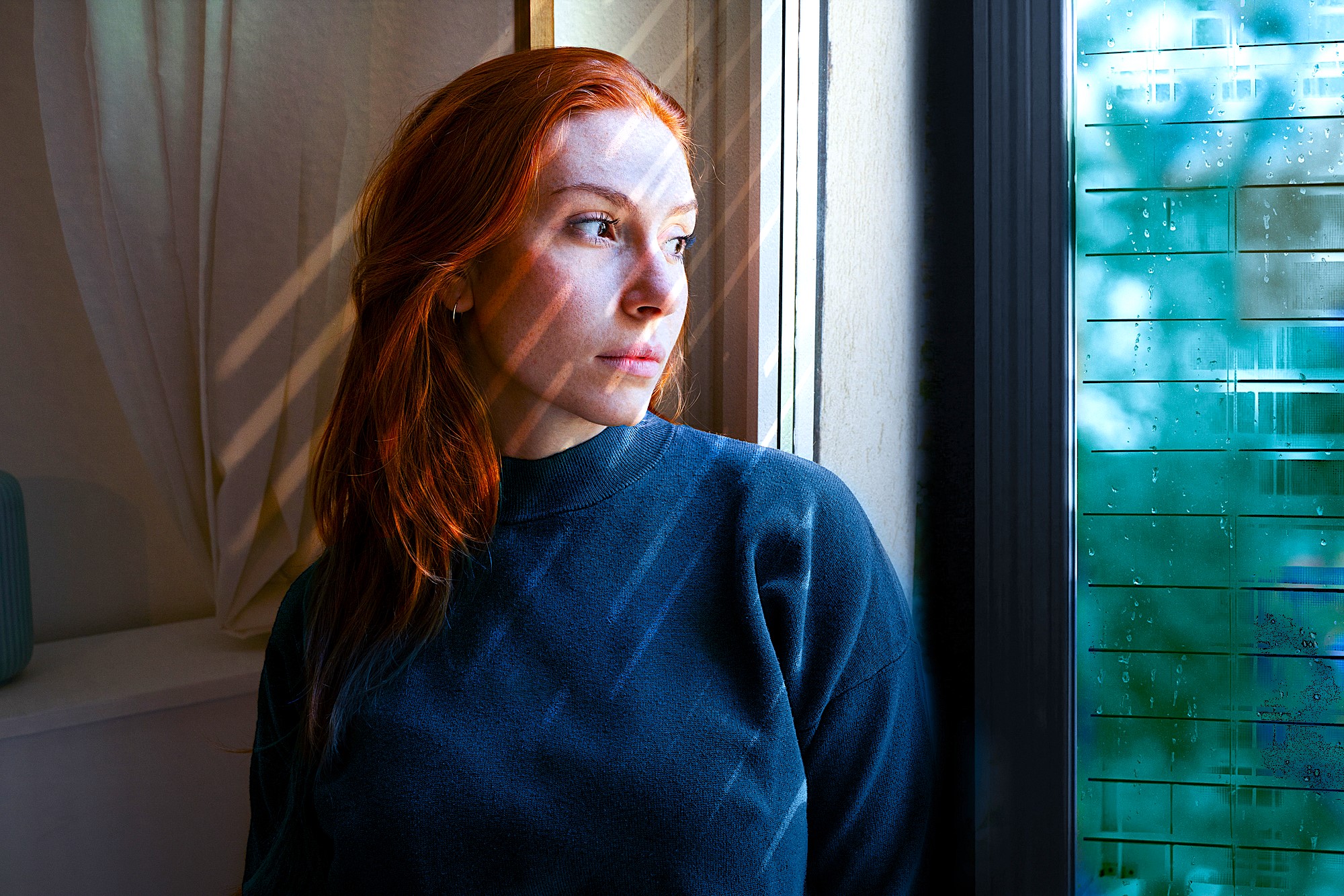 A person with long red hair wearing a dark sweater looks pensively out of a window. Sunlight filters through blinds, casting striped shadows on their face. The window reflects a mix of green foliage and sky.