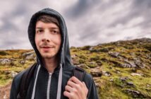 A young person in a black hoodie, with a backpack, stands outdoors against a rocky hillside and cloudy sky. The person is looking at the camera with a slight smile.