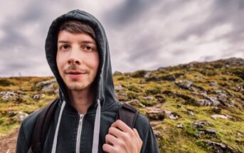 A young person in a black hoodie, with a backpack, stands outdoors against a rocky hillside and cloudy sky. The person is looking at the camera with a slight smile.