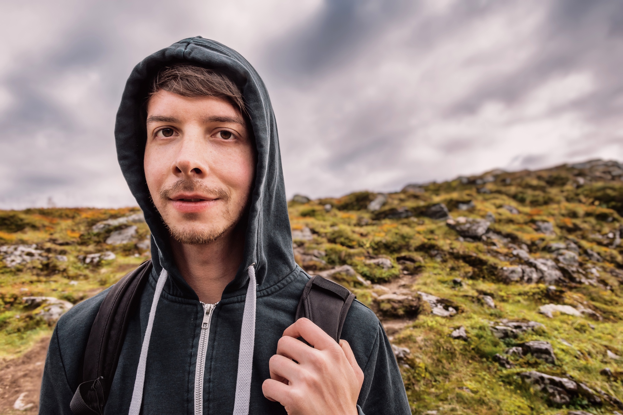 A young person in a black hoodie, with a backpack, stands outdoors against a rocky hillside and cloudy sky. The person is looking at the camera with a slight smile.