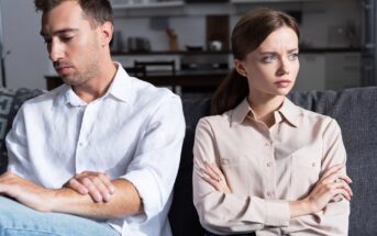 A man and woman sit on a couch, both with arms crossed and looking away from each other. The man wears a white shirt, and the woman wears a beige blouse. The setting appears to be a modern living room.