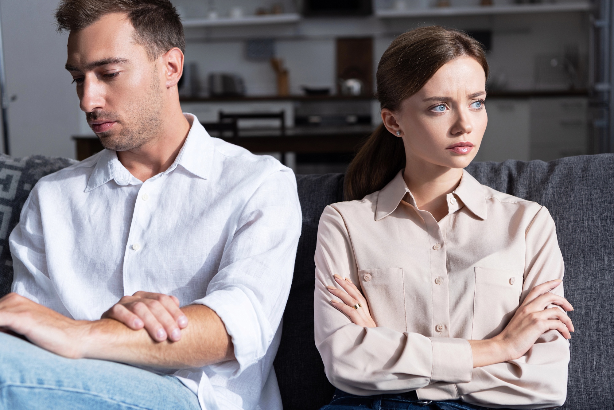 A man and woman sit on a couch, both with arms crossed and looking away from each other. The man wears a white shirt, and the woman wears a beige blouse. The setting appears to be a modern living room.