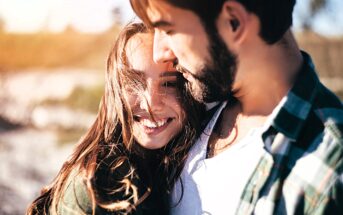 A couple standing close together outdoors. The woman is smiling and looking at the camera, while the man is slightly turned toward her. They are in a sunny, natural setting with soft, blurred background.