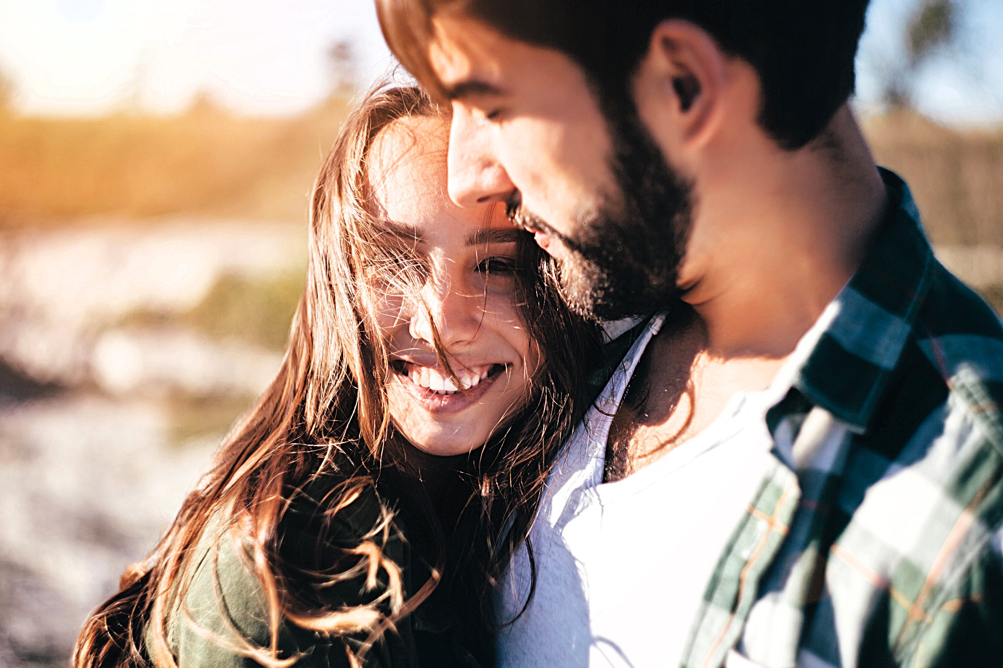 A couple standing close together outdoors. The woman is smiling and looking at the camera, while the man is slightly turned toward her. They are in a sunny, natural setting with soft, blurred background.